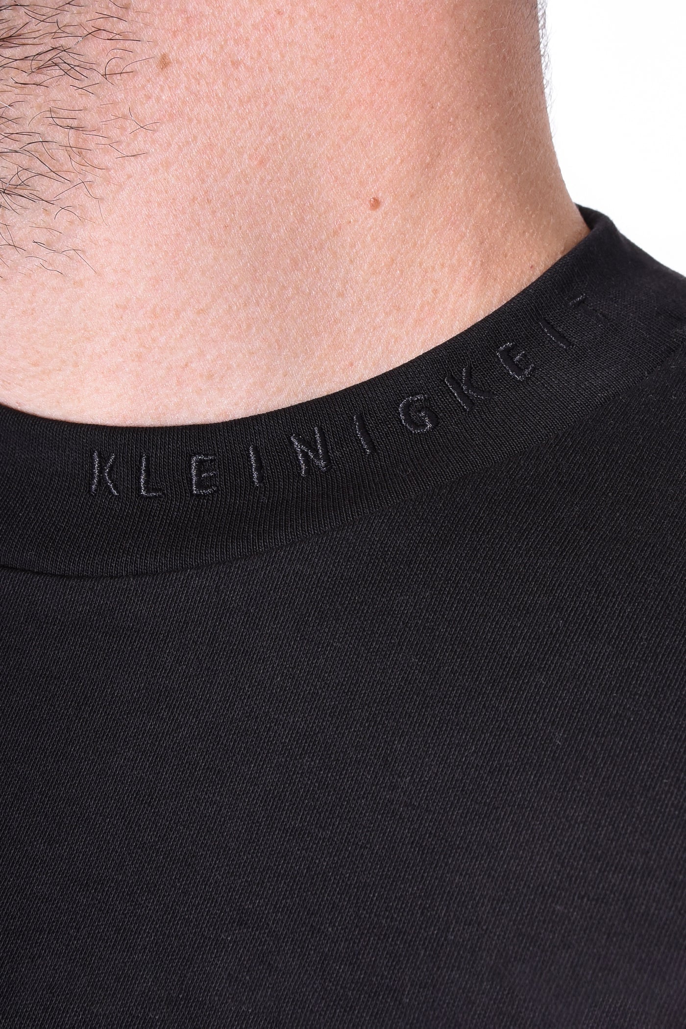 Nahaufnahme vom Kleinigkeit Bastian Feinsteiger T-Shirt mit Ton-in-Ton gesticktem Kleinigkeit Schriftzug seitlich am Kragen.