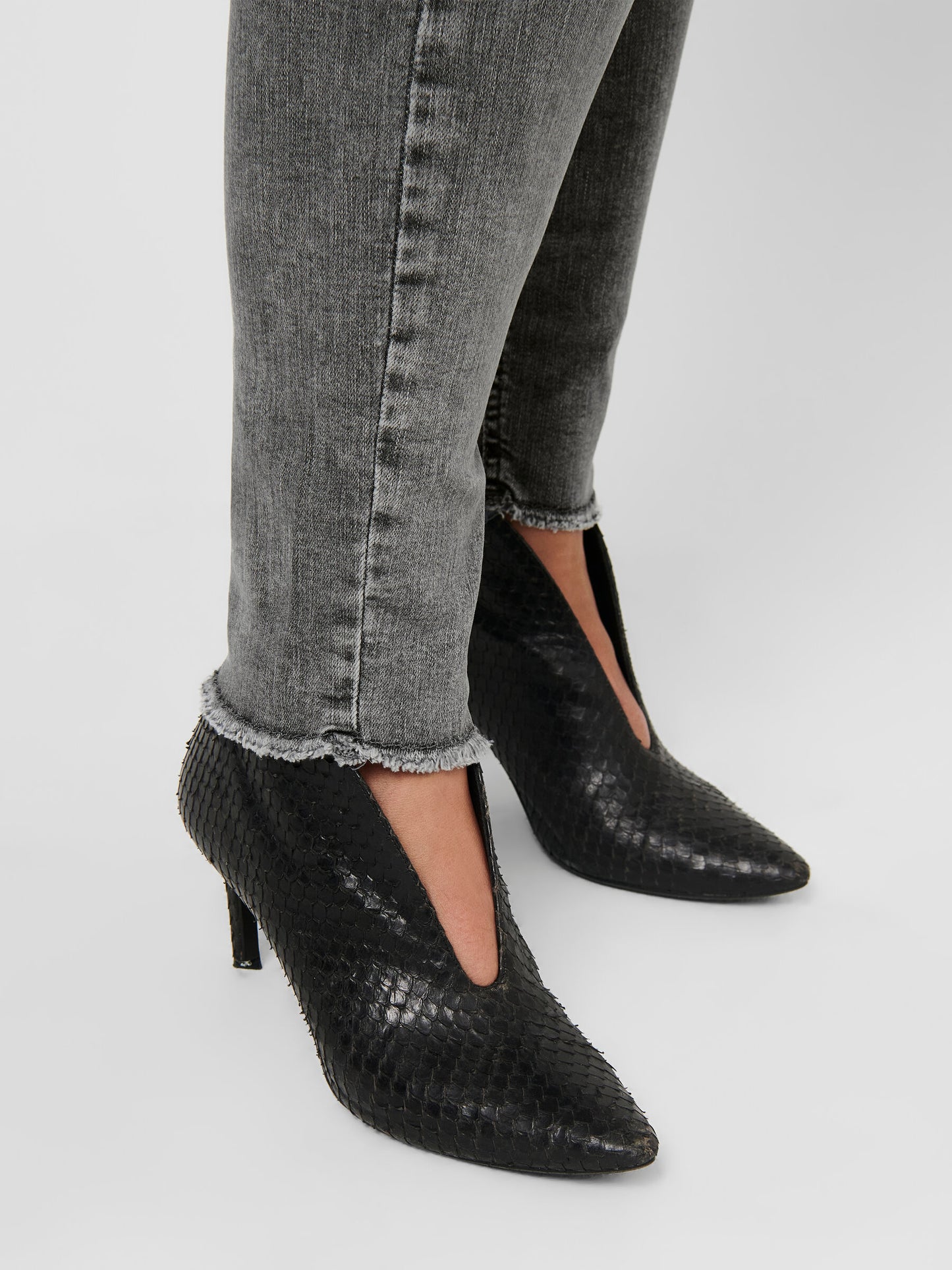 Nahaufnahme des Saums der Only Carmakoma Carwilly Skinny Grey Jeans. Zeigt den ausgefransten Beinabschluss, kombiniert mit schwarzen High Heels.