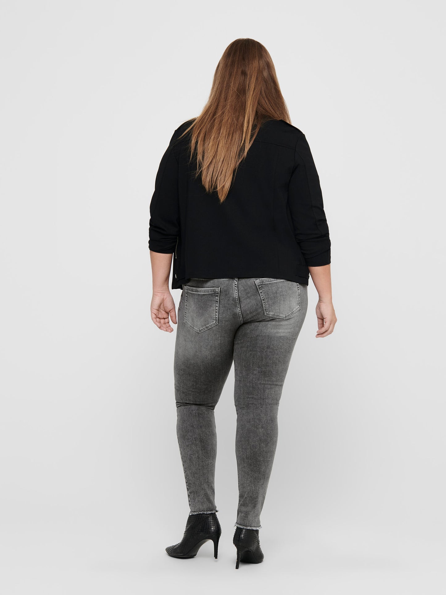 Rückansicht der Only Carmakoma Carwilly Skinny Grey Jeans, getragen von einem Model mit schwarzer Jacke und High Heels. Die graue Jeans betont die Figur und sorgt für einen modernen Look.