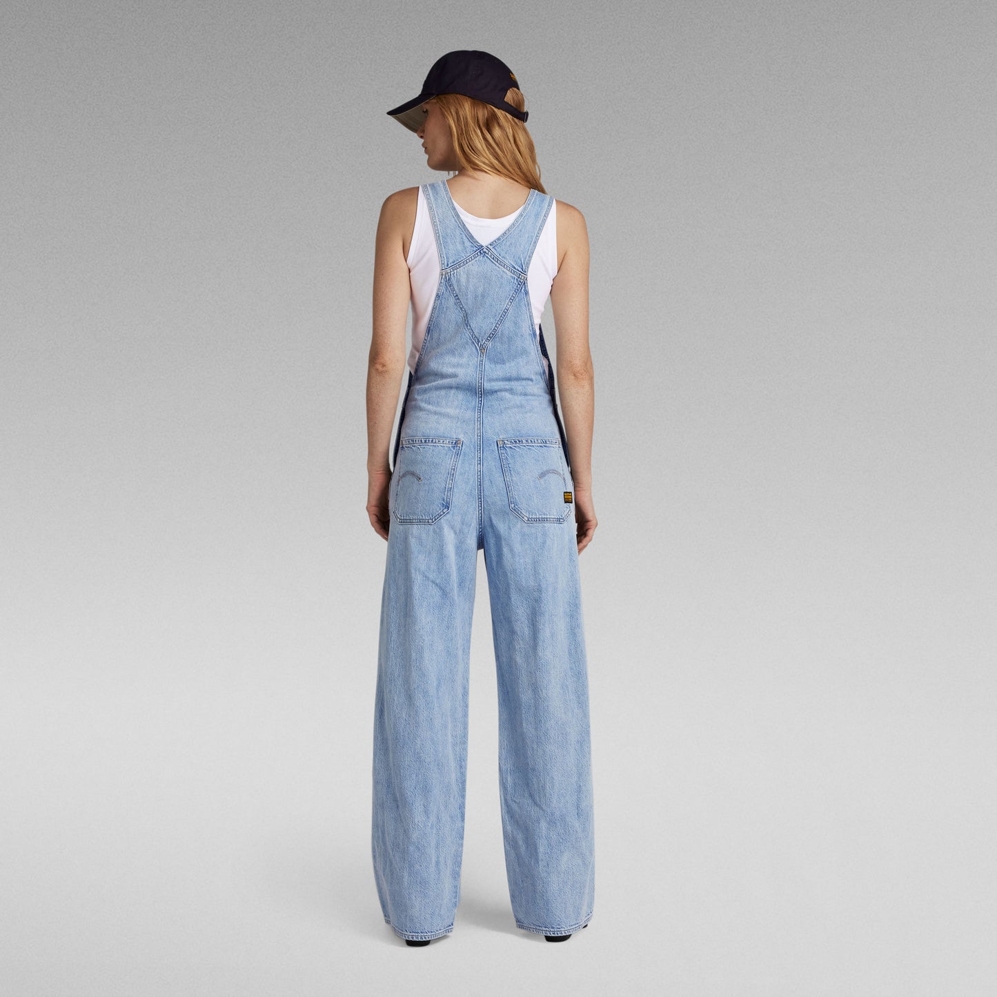 Rückansicht einer Damen Latzhose von G-Star aus hellblauem Denim mit weitem Bein, kombiniert mit einem weißen Tanktop, schwarzer Cap und schwarzen Schuhen.