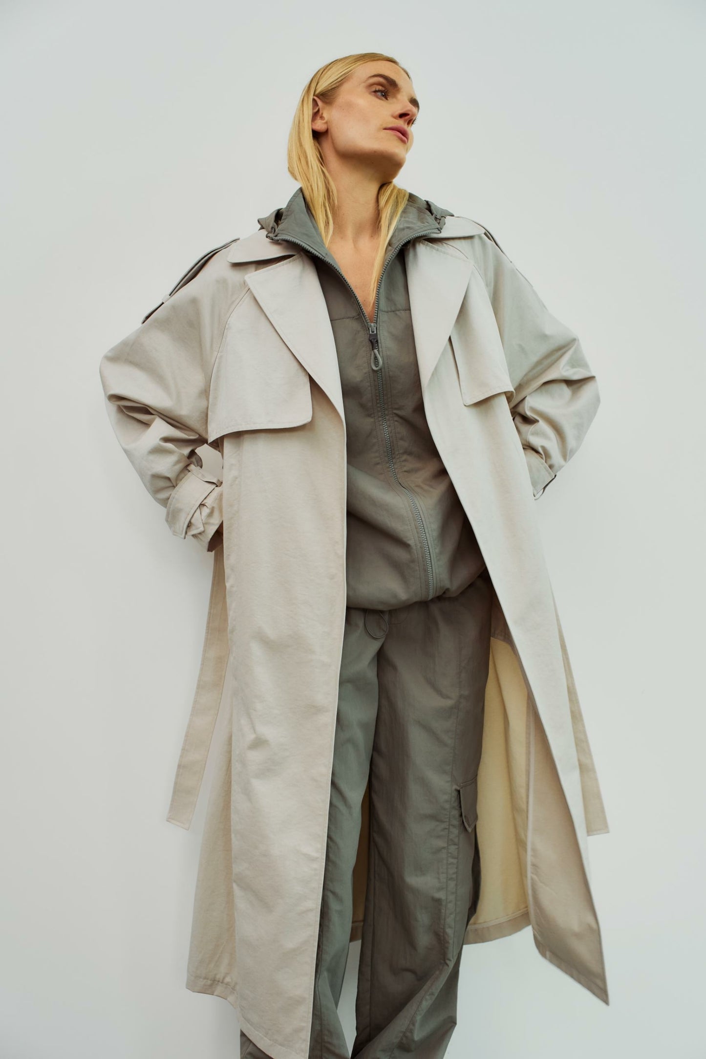 Model trägt einen beigen Trenchcoat von WithBlack über einer grauen Kapuzenjacke und einer Cargohose für einen lässigen Look.