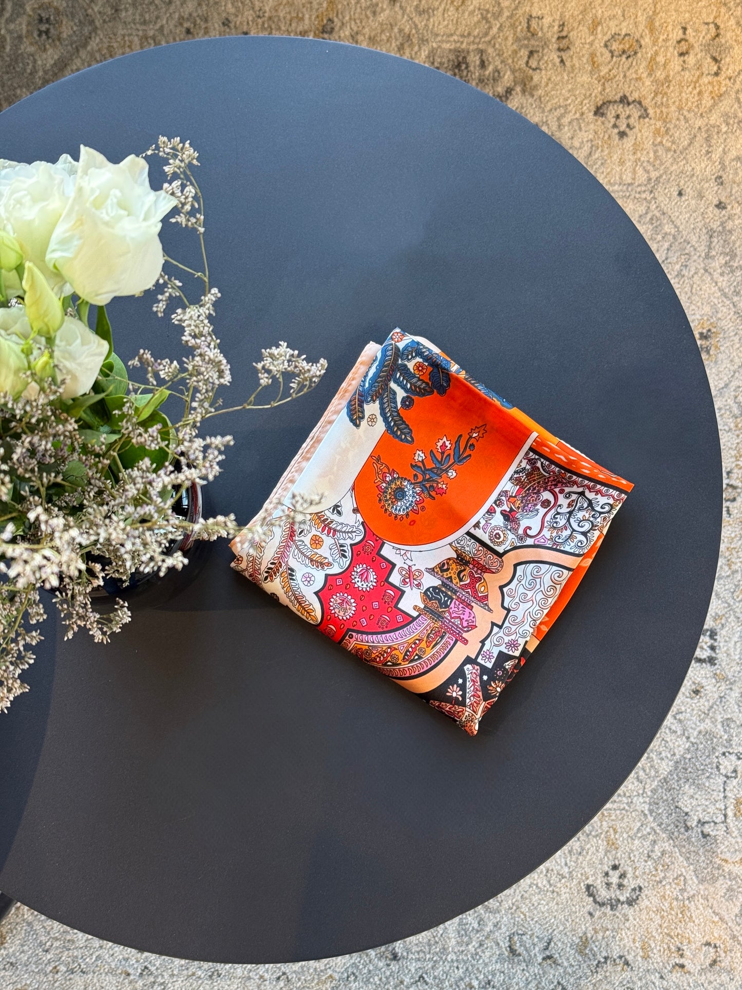 Hochwertiges Twilly Grande Carré Seidentuch mit kunstvollem Design in Orange, Rot und Weiß, edel gefaltet auf dunklem Tisch präsentiert, neben einem Blumenarrangement.