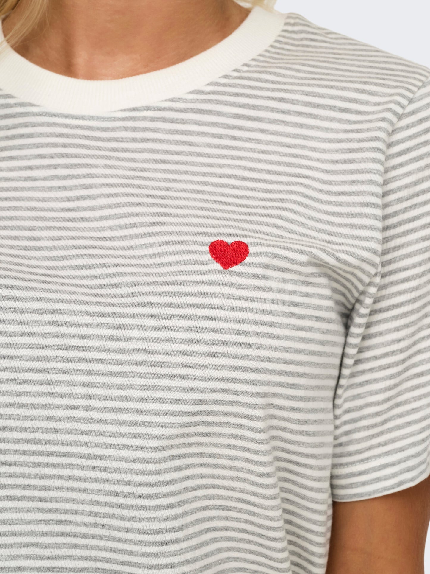 Nahaufnahme des JDY Pisa Tops in der Farbe Cloud Dancer Stripes mit rotem Herz-Detail auf der Brust. Die feinen Streifen und die liebevolle Stickerei verleihen dem Shirt eine besondere Note.