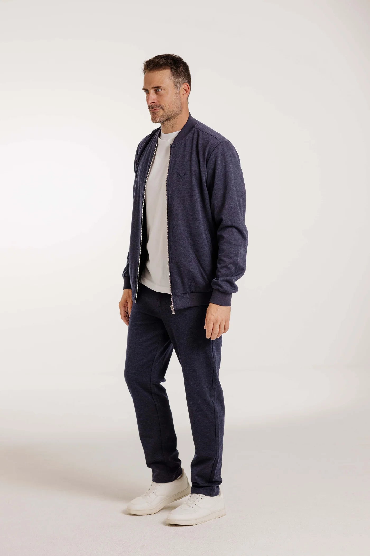 Mann trägt den Kleinigkeit Peter Maffein Blouson Navy Blue mit passender Hose und weißen Sneakern in einer neutralen Studioaufnahme.