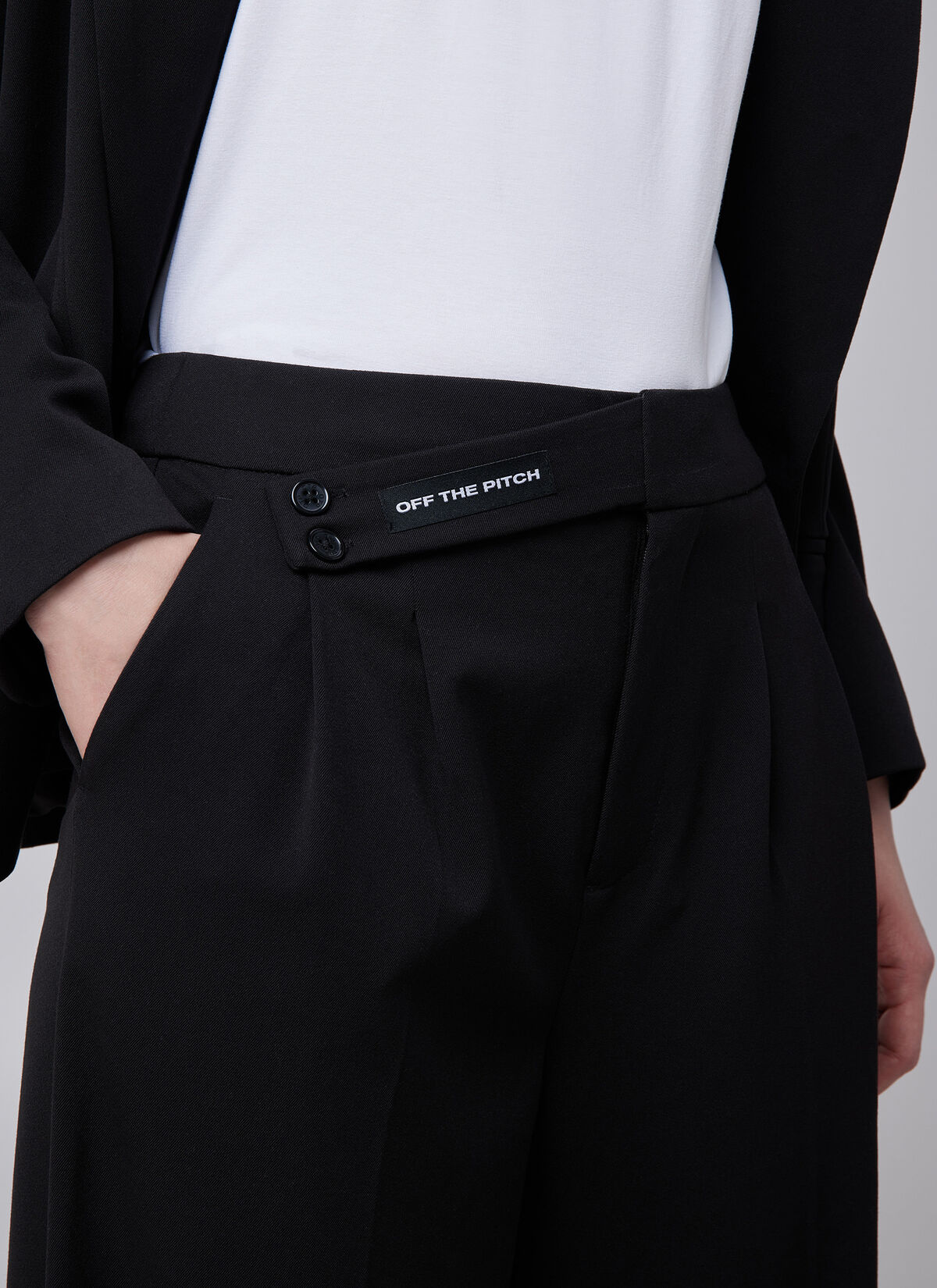 Off the Pitch Pleated Gaia Pants Black – Detailaufnahme des Bunds mit sichtbarem Logo-Label „Off the Pitch“ und doppeltem Knopfverschluss.