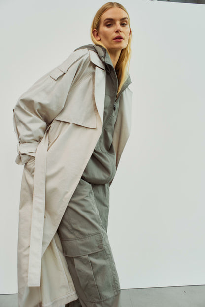 Model posiert in einem beigen Trenchcoat von WithBlack mit lockerer Silhouette, getragen über einer sportlichen grauen Kapuzenjacke.