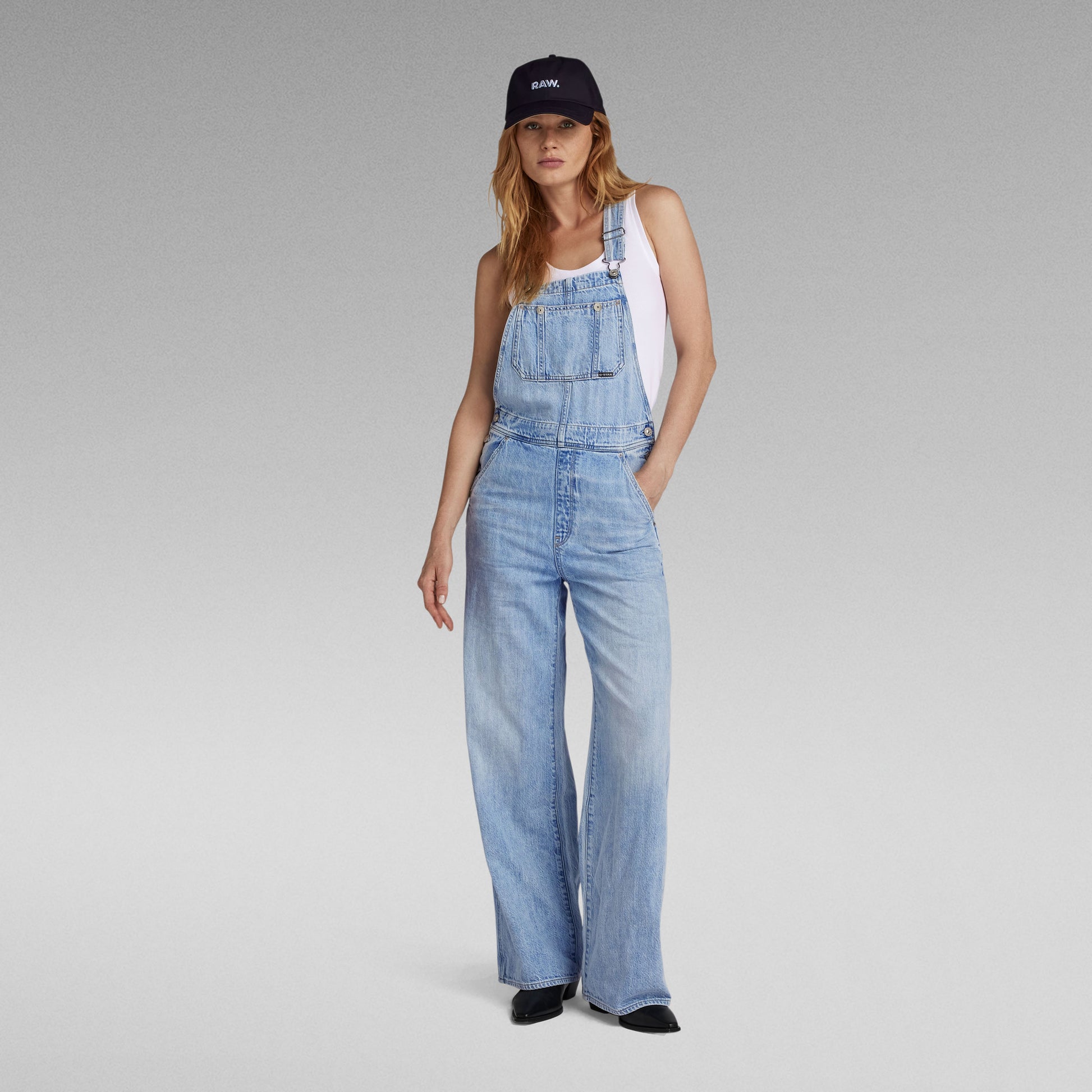 Frontansicht einer Damen Latzhose von G-Star aus hellblauem Denim mit weitem Bein, kombiniert mit einem weißen Tanktop, schwarzer Cap und schwarzen Schuhen.