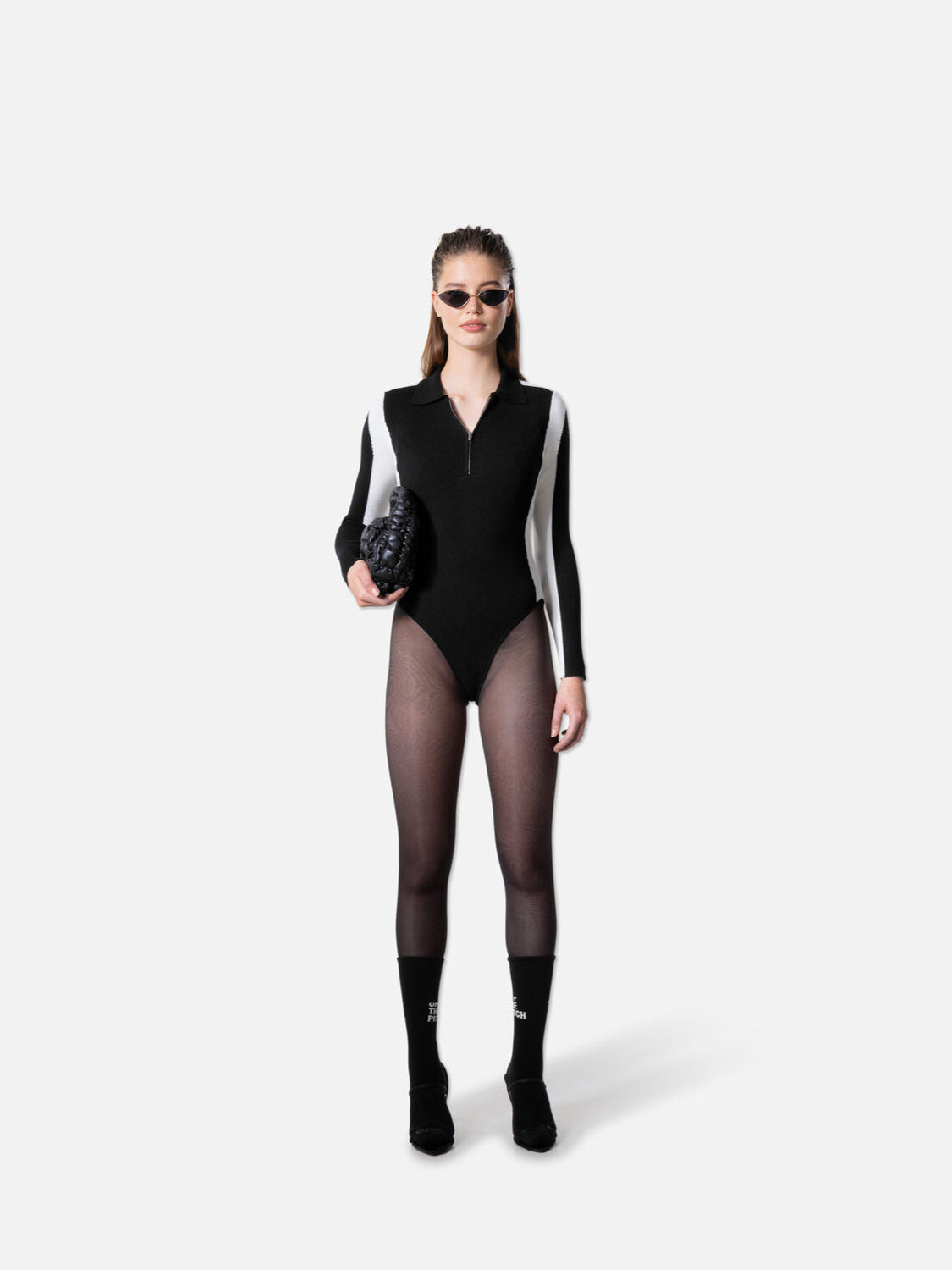 Model trägt einen schwarz-weißen langärmligen Body mit Reißverschluss, kombiniert mit transparenten Strumpfhosen, schwarzen Sonnenbrillen, einer strukturierten schwarzen Clutch und schwarzen Socken mit weißem Schriftzug, dazu schwarze Schuhe, vor einem weißen Hintergrund fotografiert.