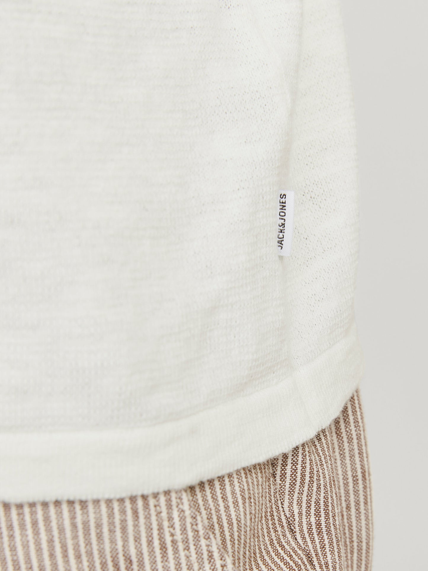 Detailansicht des Saums mit Label des Jack & Jones Summer Knit Crew Neck Pullovers in Weiß, kombiniert mit braun-weiß gestreifter Hose.