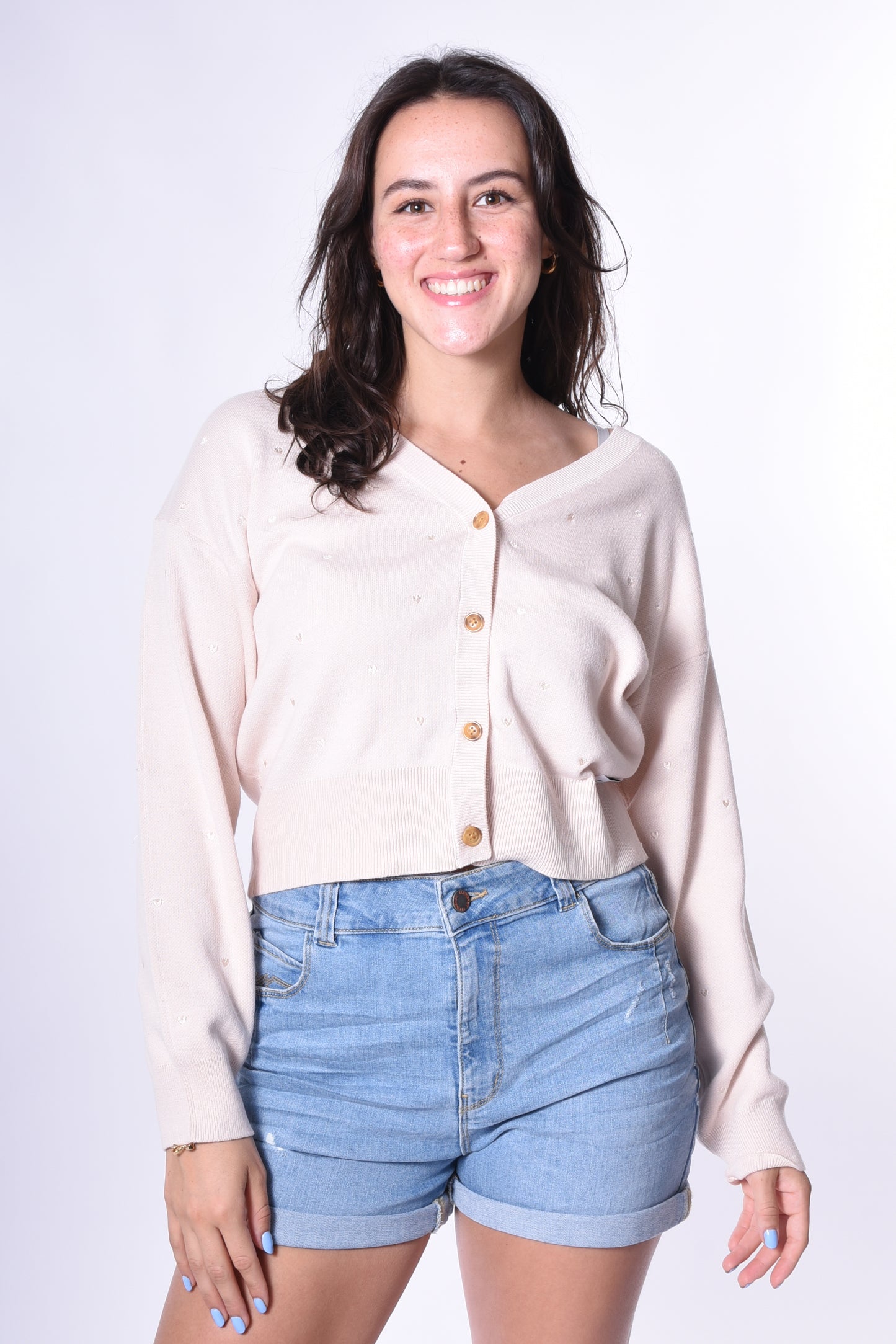 Junge Frau trägt den Kleinigkeit Désirée Strick-Cardigan in Beige mit kleinen eingestickten Herzen und Holzknöpfen, kombiniert mit blauen Jeans-Shorts. Ein lässig-eleganter Look für jede Gelegenheit.