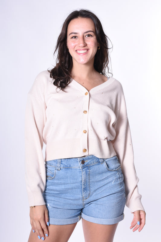 Junge Frau trägt den Kleinigkeit Désirée Strick-Cardigan in Beige mit kleinen eingestickten Herzen und Holzknöpfen, kombiniert mit blauen Jeans-Shorts. Ein lässig-eleganter Look für jede Gelegenheit.