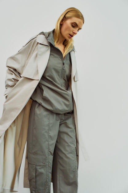 Model präsentiert einen beigen Trenchcoat von WithBlack mit Bindegürtel kombiniert mit grauer Kapuzenjacke und Cargohose.
