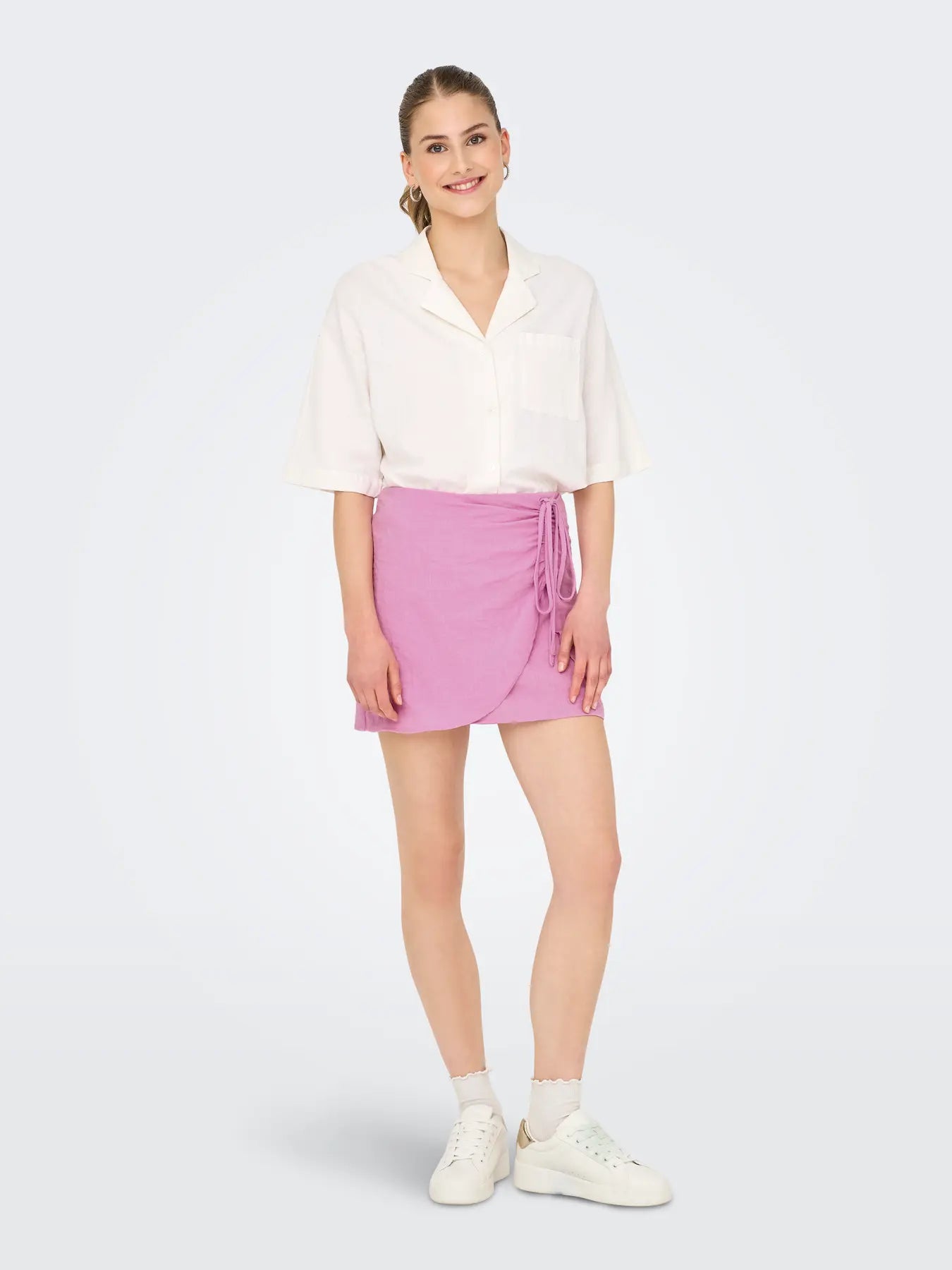 Modell trägt den Only Emmia Life Skirt in Crocus, Minirock mit Wickeloptik und Bindeband in Flieder, kombiniert mit weißer Bluse und Sneakern.