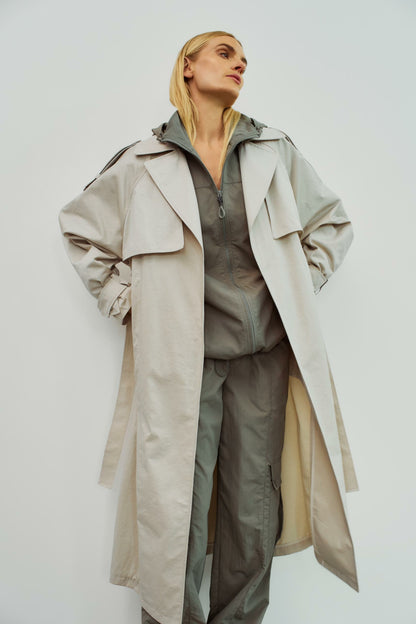 Model trägt einen beigen Trenchcoat von WithBlack über einer grauen Kapuzenjacke und einer Cargohose für einen lässigen Look.