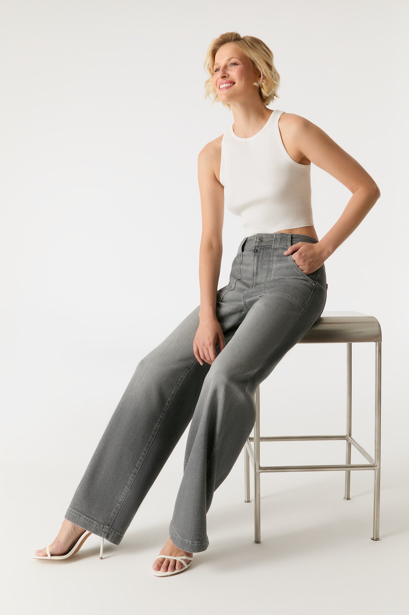 COJ Lulu Wide Leg Jeans in Grey Vintage, getragen von einem sitzenden Model auf einem Hocker, kombiniert mit einem weißen Crop-Top.