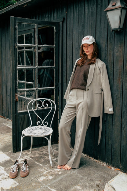 Frau trägt den Colourful Rebel Izzy Belted Pinstripe Blazer - Beige lässig kombiniert mit Kappe und Strickpullover vor dunkler Holzwand