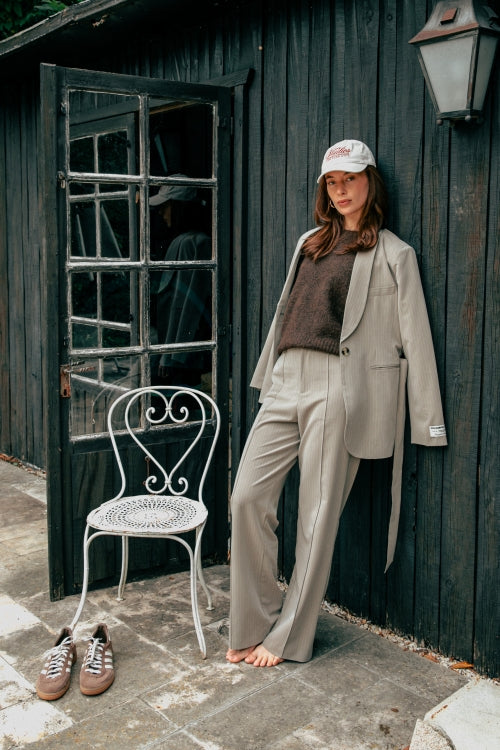 Frau trägt den Colourful Rebel Izzy Belted Pinstripe Blazer - Beige lässig kombiniert mit Kappe und Strickpullover vor dunkler Holzwand