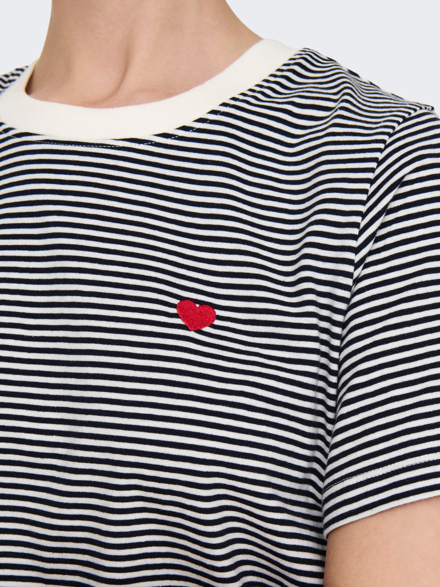 Nahaufnahme des JDY Pisa Tops in der Farbe Cloud Dancer Sky Captain Stripes mit rotem Herz-Detail auf der Brust. Die feinen Streifen und das kleine gestickte Herz setzen dezente Akzente.