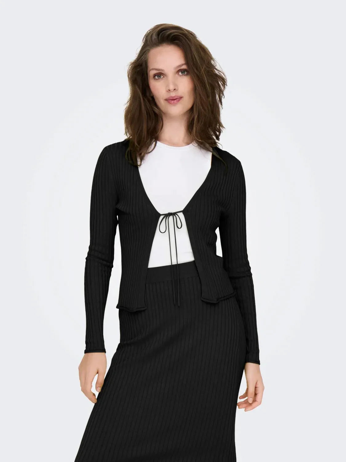 Model trägt den Only Mila Cardigan Black, einen schwarzen Rippstrick-Cardigan mit Bindeband vorne, kombiniert mit einem weißen Oberteil und einem passenden schwarzen Rock.