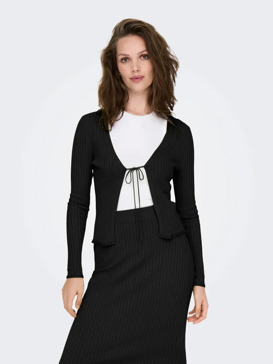 Model trägt den Only Mila Cardigan Black, einen schwarzen Rippstrick-Cardigan mit Bindeband vorne, kombiniert mit einem weißen Oberteil und einem passenden schwarzen Rock.